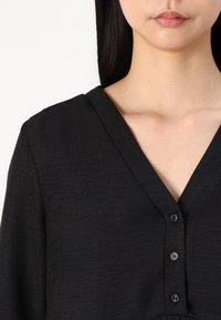 Black v-neck top with a textured fabric. Features five buttons down the front, with a smooth finish and subtle sheen.