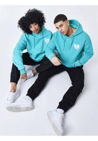 Sudaderas turquesas con gráficos de corazones, combinadas con pantalones de chándal negros y sneakers blancos. El diseño incluye capuchas con cordón y un corte relajado.
