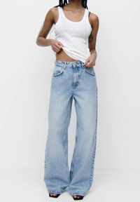 Jeans anchos de color azul claro con cintura alta, cinco bolsillos y costuras en contraste, combinados con una camiseta ajustada blanca.