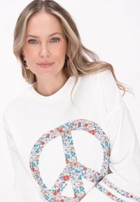 Vit sweatshirt med ett stort fredssymbol dekorerat med färgglada blommönster. Slät yta med ribbad halsringning och ärmslut.