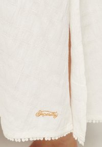Ropa de tela blanca texturizada con un patrón tejido, un dobladillo en forma de ondas con pompones y un bordado dorado del nombre de la marca.