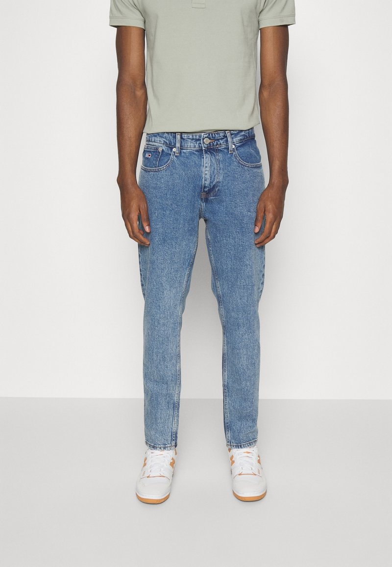 Světlomodré džíny z denimu s úzkým střihem, pěti kapsami a mírným vyblednutím. Kombinovány s bílými teniskami a světle zeleným polo tričkem.