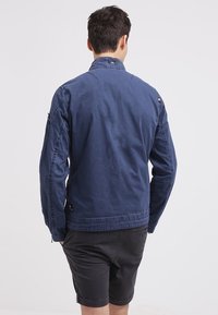 Mörkblå jacka med ståkrage, långa ärmar, sidofickor och dragkedja fram. Tyget ser slätt ut med minimal textur.