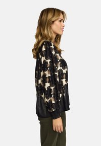 Kvinne med bølgete blondt hår iført en svart og beige blomstret langermet bluse og olivengrønne bukser, stående i profil mot en hvit bakgrunn.