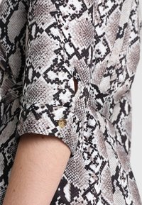 Gros plan d'un bras portant une manche de chemise roulée à motif serpent avec un petit bouton marron sur le poignet.