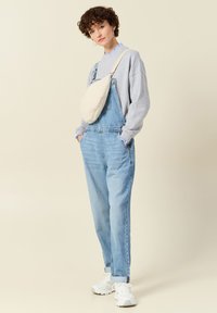 Overoles de mezclilla de un azul claro con un corte holgado, combinados con una sudadera gris y un bolso de hombro color crema. El conjunto cuenta con zapatillas blancas.