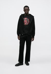 Black zip-up hoodie with a front pocket, worn over a black t-shirt featuring a red Deadpool graphic, paired with loose black pants and black shoes.