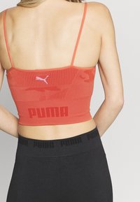 Rød atletisk crop-top med tynde stropper, der har et tekstureret design og et fremtrædende sort "PUMA" logo ved taljen. Matchet med sorte leggings.