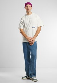 Tricou alb cu mânecă scurtă, având logo-ul "another" pe piept, asortat cu blugi denimi largi de culoare albastră și adidași cu accente verzi.