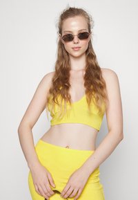 Conjunto de top e saia de toalha de algodão amarelo com design de decote em V. Modelo usa óculos de sol finos e coloridos e tem cabelo longo e ondulado.