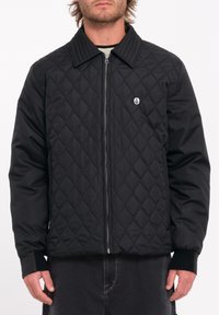 Chaqueta negra acolchada con cremallera en la parte delantera, cuello y dobladillo acanalados. Presenta un pequeño logo blanco en el pecho. El material parece suave y acolchado.