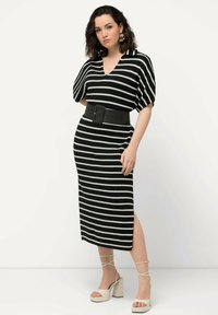 Robe tricot rayée noir et blanc avec un col en V, des manches courtes et une ceinture noire. La modèle porte des sandales à talons blocs beige.
