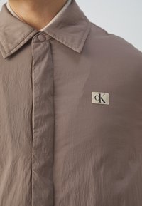 Brown button-up shirt with a textured finish, featuring a pointed collar and a cream-colored CK logo patch on the chest.