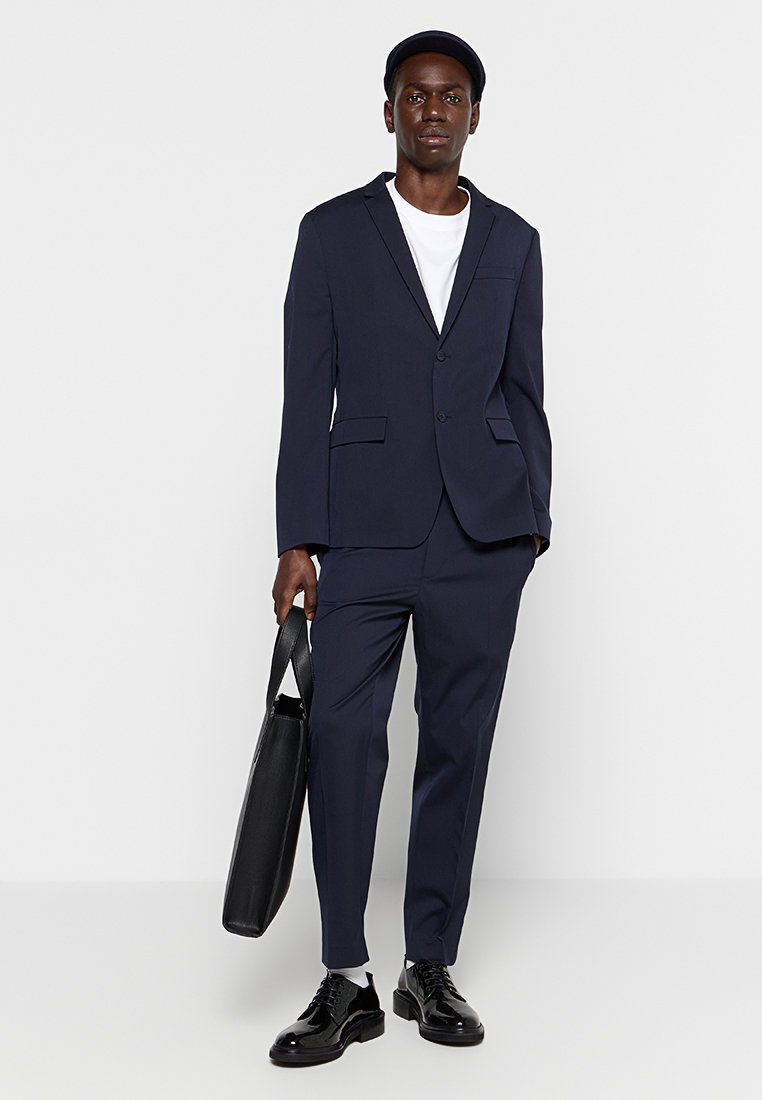 Costume marine à la coupe élégante, comprenant un blazer à bouton unique et un pantalon sur mesure. Assorti à un sac noir et des chaussures noires brillantes.