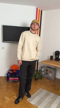Sweatshirt bege com logotipo, calças cargo pretas e sapatilhas pretas. O fundo apresenta riscas coloridas na parede e elementos de decoração moderna.