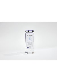 Flacon de shampooing Kérastase Blond Absolu Bain Lumière avec bouchon argenté sur support transparent, entouré de mousse, fond blanc.