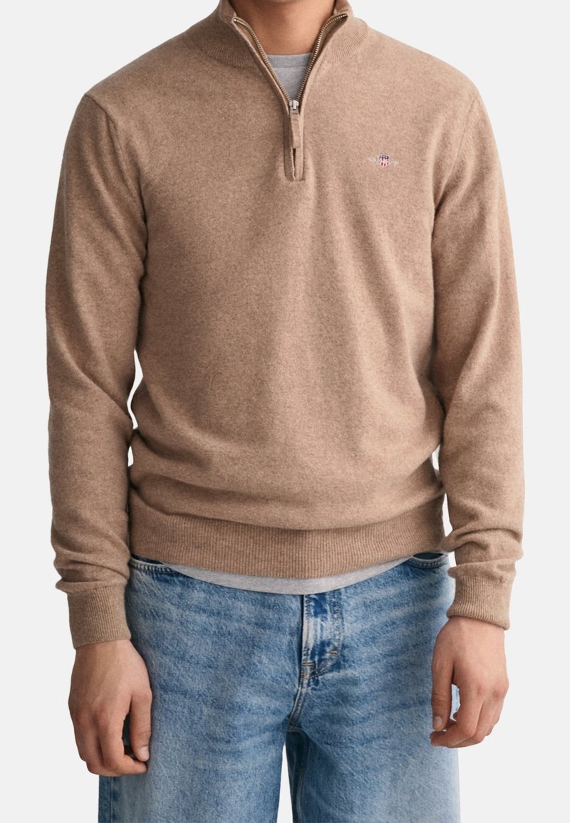 Brauner Reißverschluss-Pullover aus weichem Strickmaterial, mit hohem Kragen und gerippten Bündchen. Getragen mit hellblauen Jeans.