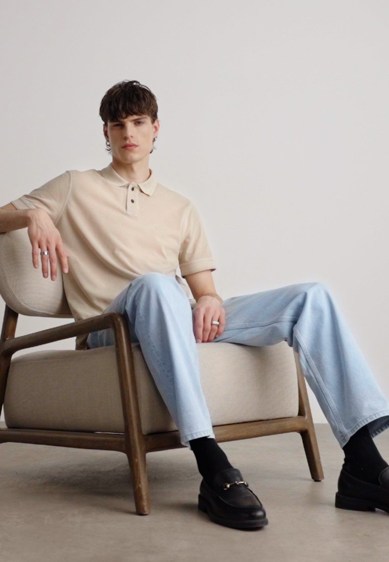 Chemise polo beige à manches courtes, jean droit bleu clair, mocassins noirs avec accents dorés, assis sur une chaise de couleur claire.