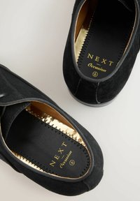 Ein Paar schwarze Wildleder-Slipper mit goldverzierten Innensohlen, markiert mit "NEXT Occasion", auf einer hellen Fläche präsentiert.