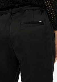 Zwarte broek van gladde stof met een achterzak met een elegante zwarte bies en een klein zilveren label voor detailing.