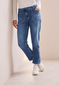Jeans en denim bleu clair avec une coupe décontractée, dotés de poches avant doubles et d'une taille à cordon. Associés à des baskets blanches.