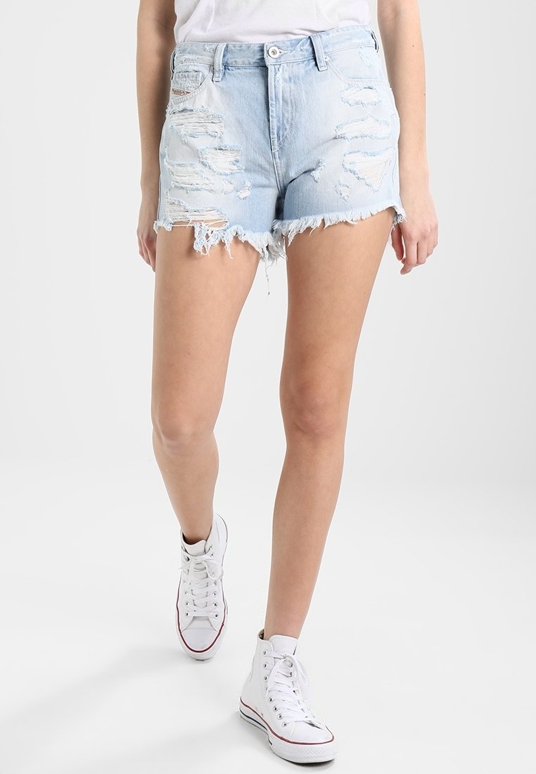 Femme marchant en short en jean déchiré bleu clair et baskets montantes blanches sur un fond blanc uni.