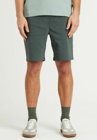 Pantaloni shorts verde scuro realizzati in tessuto leggero con una texture liscia. Design classico, lunghezza fino al ginocchio con orlo ripiegato, abbinati a sneaker bianche.