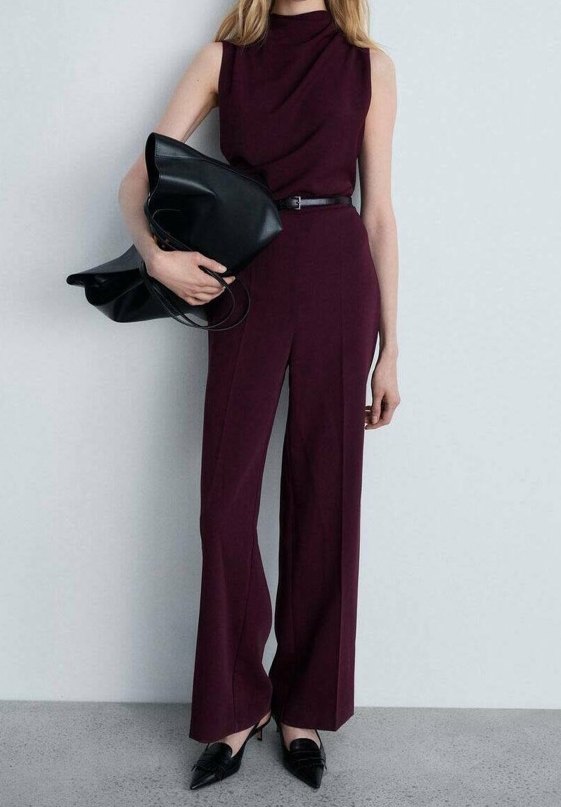 Femme en combinaison bordeaux sans manches avec ceinture noire, tenant un grand sac en cuir noir et portant des chaussures noires pointues.