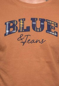 T-shirt en coton marron avec "BLUE" en grandes lettres en plaid bleu marine avec des accents jaunes et "& Jeans" en cursive en dessous. Texture lisse.