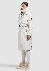 Bílý a béžový hybridní kabát s vlněnou texturou, nadýchanými rukávy, vysokým límcem, detailem s páskem a kapsami na zip. Kombinován s béžovými botami.
