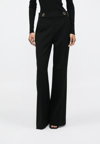 Mulher a usar calças largas de cintura alta pretas com botões dourados na cintura e sapatos pretos de salto aberto, de pé contra um fundo plain.