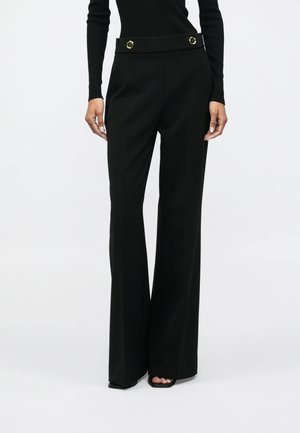 Woman wearing high-waisted black wide-leg pants with gold buttons at the waistband and black open-toe shoes, standing against plain background.