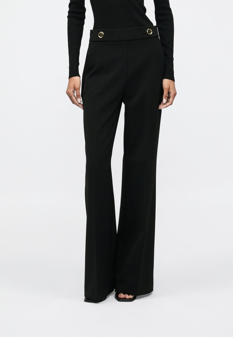 Mulher a usar calças largas de cintura alta pretas com botões dourados na cintura e sapatos pretos de salto aberto, de pé contra um fundo plain.