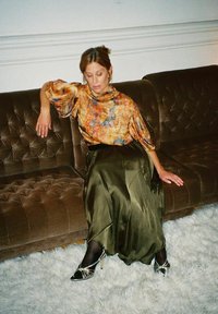 Tryckt blus i varma färger, olivgrön satinskirt och svarta klackar med silverdetaljer, sittande på en brun stoppad soffa med vit matta.