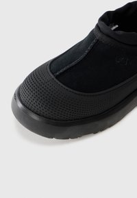 Black UGG slip-on shoe with textured rubber toe cap and thick sole, featuring suede upper and padded collar.