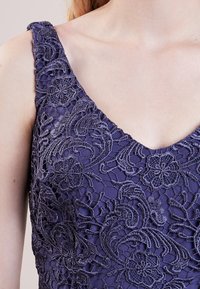 Lila klänning med intrikata blommönster i spets med scalloped kant och breda axelband, som framhäver en strukturerad mönstring och V-ringning.