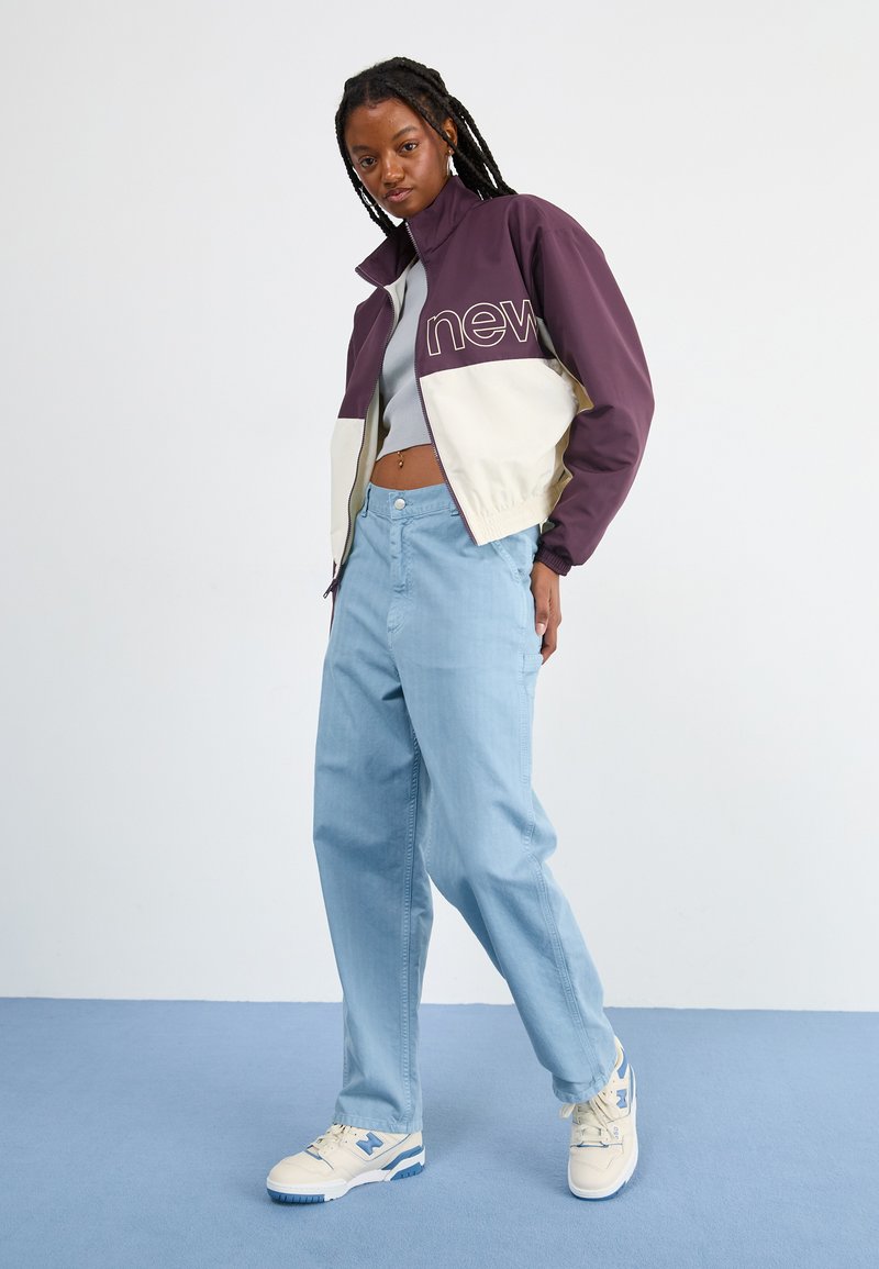 Giacca viola e beige con testo "nuovo", pantaloni a gamba larga azzurri e scarpe da ginnastica bianche con accenti blu. Tessuti lisci e vestibilità comoda.