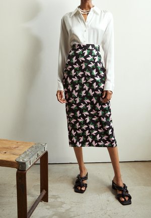 Sort nederdel med pink og grøn blomsterdesign, parret med en hvid bluse. Modellen har sorte klodsede sandaler på og står på et beton gulv.