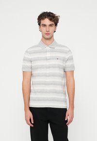 Camisa polo a rayas gris y blanca con cuello estándar y mangas cortas. Cuenta con un logotipo en el pecho y una tela texturizada. Combinada con pantalones negros.