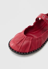 Roter Lederschuh mit plissiertem Design, abgerundeter Spitze und schwarzer Sohle mit Nietenakzenten. Verstellbarer Riemen mit dekorativer Hardware.