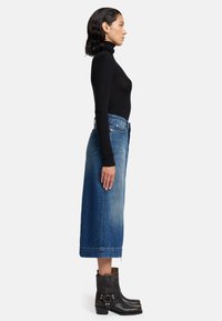 Haut noir à col roulé côtelé associé à une jupe midi en denim bleu. La tenue comprend des bottes noires à talon avec des accents métalliques.