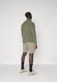 Veste en polaire verte, short cargo beige, chaussettes noires et baskets gris clair. Texture lisse et design décontracté, avec des poches sur le short.
