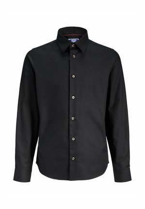 Junge trägt schwarzen Kapuzenpullover mit Reißverschluss, schwarzes Hemd mit Knöpfen, graue Jeans und schwarze Sneakers, steht vor weißem Hintergrund.