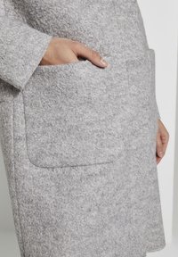 Grå texturerad kappa med en synlig ficka. Materialet verkar mjukt och något fluffigt, med en skräddarsydd, rak design.