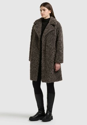 Femme portant un long manteau en fausse fourrure texturée gris foncé, des leggings noirs, un col roulé noir et des bottes noires montantes jusqu'aux genoux, debout sur un fond uni.