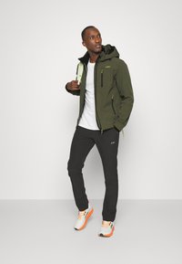 Grønne softshell-jakke med hætte, med sorte detaljer og lynlåse, parret med sorte bukser og hvid-orange sneakers.