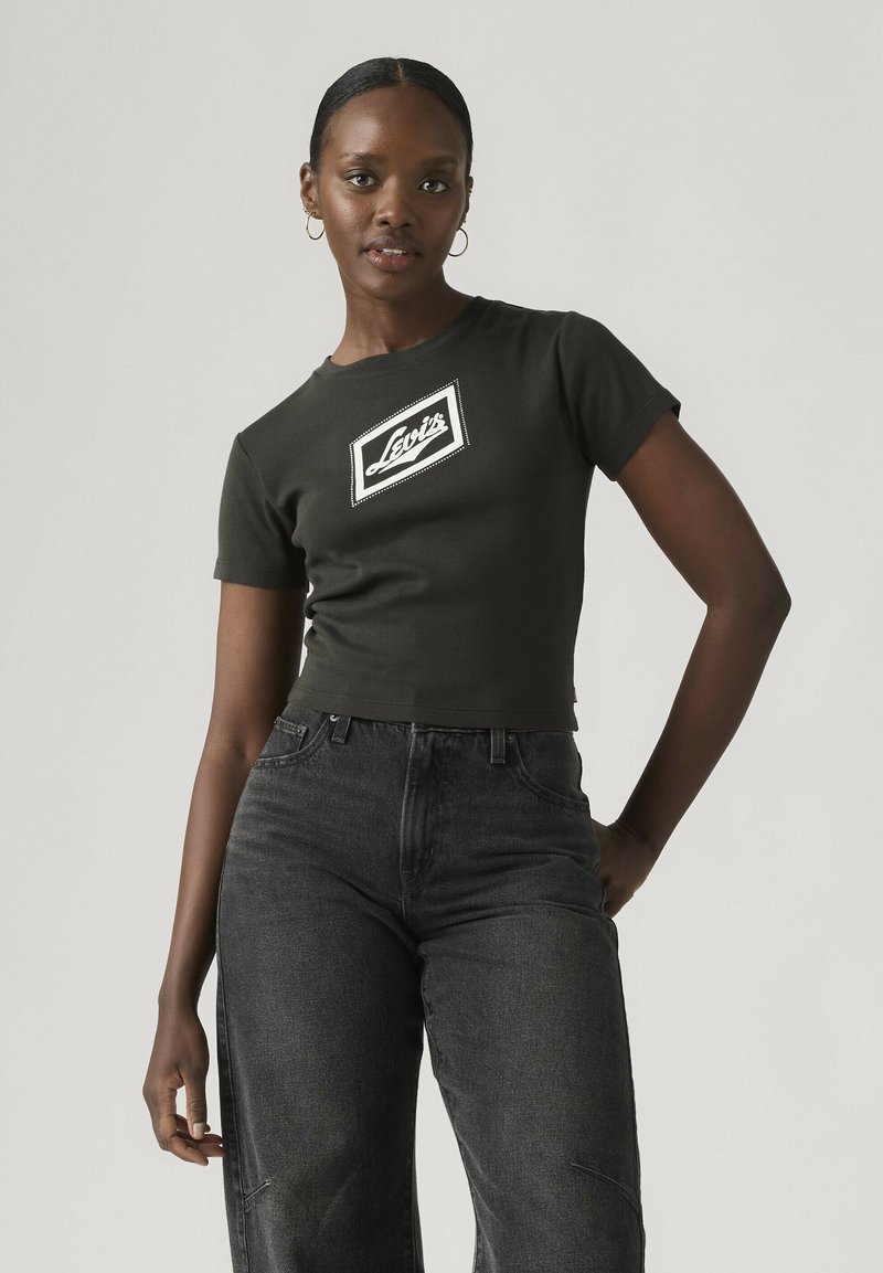 Jonge vrouw met hoepeloorbellen, gekleed in een zwart, cropped Levi’s T-shirt en donkere, hooggetailleerde spijkerbroek, staand tegen een effen achtergrond.