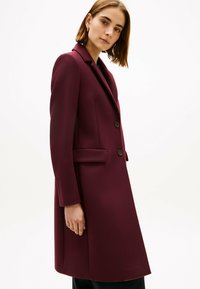 Bordeauxrode jas van een wolmix met een getailleerd ontwerp, een reverskraag met inkeping, twee knopen en zijzakken. Soepele textuur, silhouet tot op de knieën.