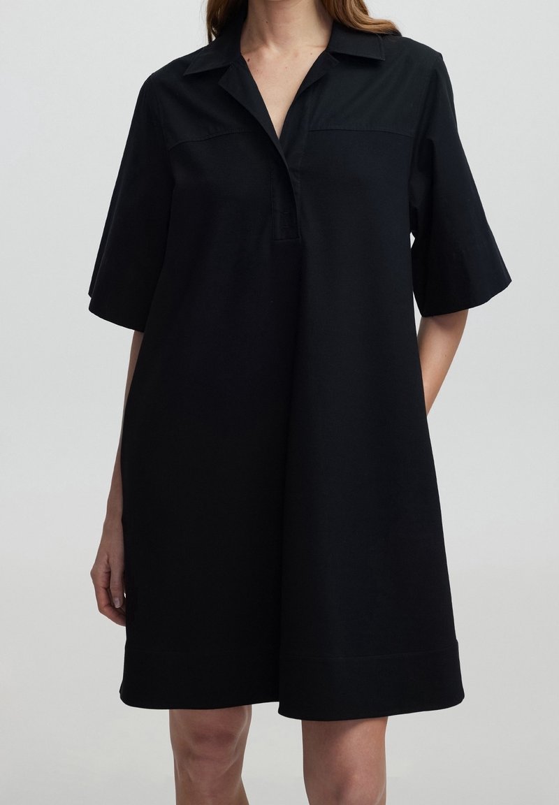 Femme portant une robe noire ample longueur genou avec des manches courtes et un col avec un petit plastron à boutons.