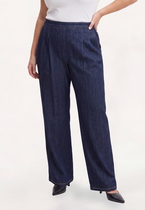 Frau trägt dunkelblaue Jeans mit weitem Bein und schwarze spitze Absätze, mit einer Hand auf der Hüfte und einem weißen Oberteil.